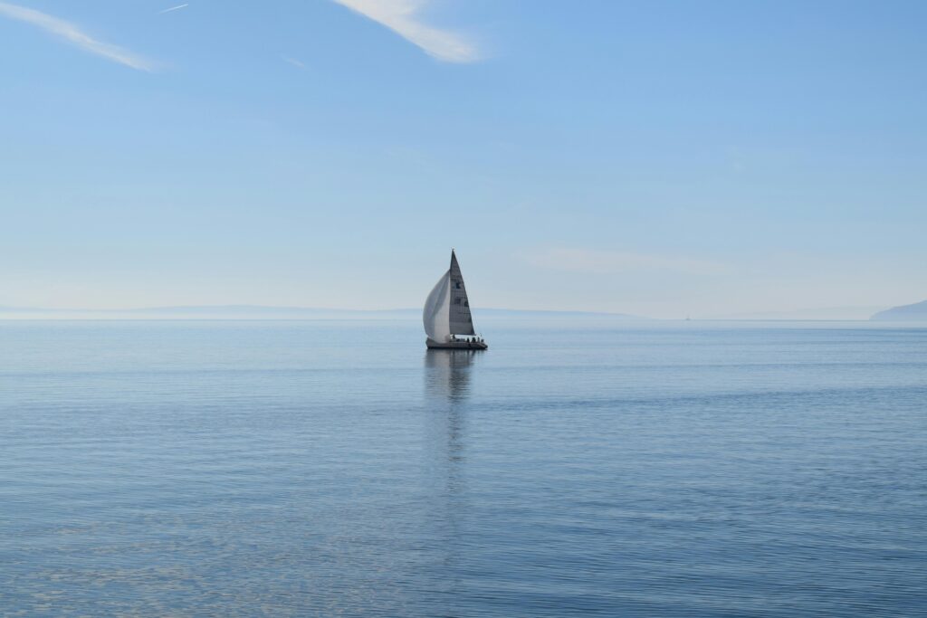 A lone sailboat glides quietly across the calm ocean under a clear blue sky.