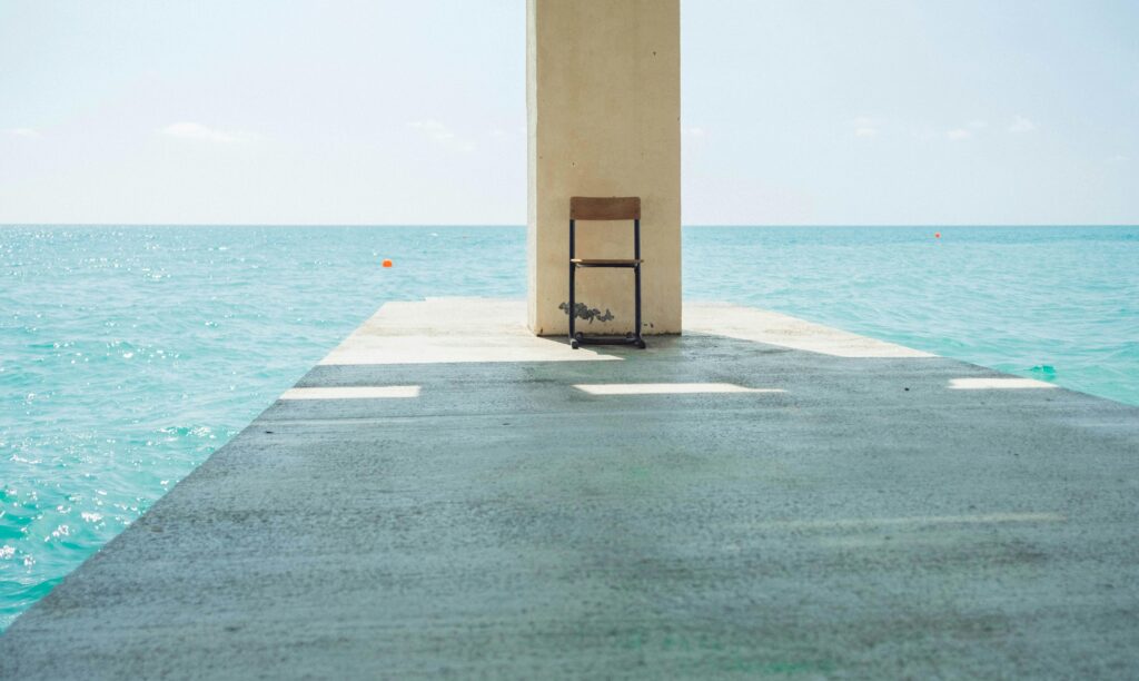 A single chair sits on a pier with a clear view of the calm ocean under a bright sky.