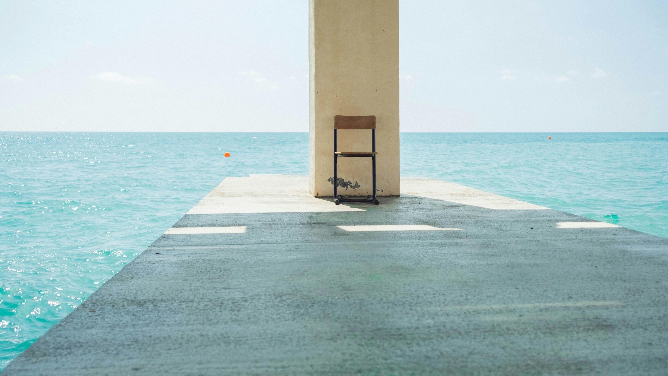 A single chair sits on a pier with a clear view of the calm ocean under a bright sky.